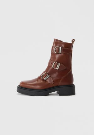 Bota de tornozelo em couro castanho com três fivelas prateadas ajustáveis, bico arredondado, costura mínima e sola plataforma preta robusta.