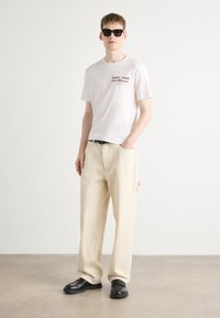 Hvid t-shirt med GANT 1949 grafik, beige vide bukser, sort bælte og sorte sko; modellen bærer sorte solbriller; minimal baggrund.