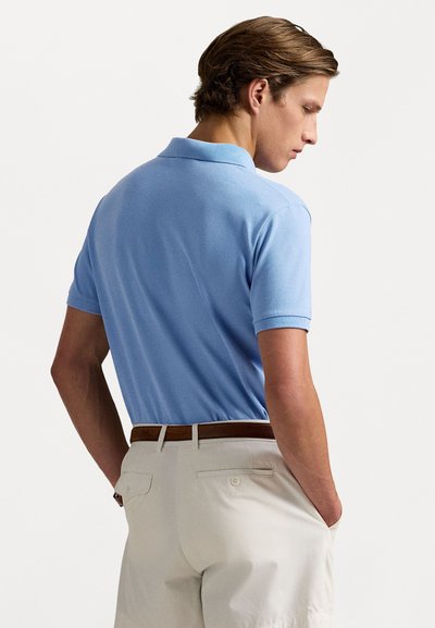 Cămașă polo de culoare albastru deschis cu mâneci scurte, guler ribflat, textură netedă, asociată cu pantaloni bej și o curea maro, văzută din spate.
