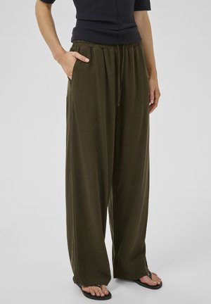 Naine, kes kannab oliivrohelisi laiade säärtega pükse elastse vöökohaga ja nööriga, musta varrukateta topi ning musti plätusid.