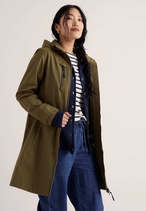 Donna con lunga treccia che indossa un cappotto con cappuccio verde oliva, cardigan blu navy, maglia a righe e pantaloni di jeans blu larghi su sfondo neutro.