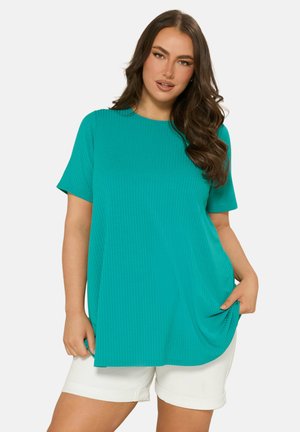 Vrouw met lang bruin haar, gekleed in een turquoise geribd kort mouwtopje en witte korte broek, houdt aan één kant de zoom van het topje vast.