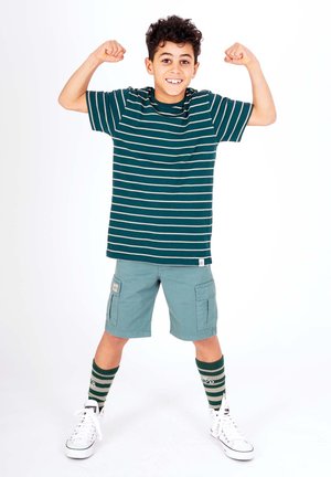 Stribet grøn t-shirt, korte lyseblå cargo shorts, grønne og hvide stribede sokker, og hvide sneakers. Dreng der flexer sine arme og smiler.