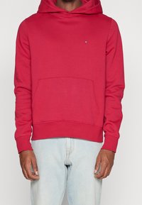 Hombre que lleva una sudadera roja con bolsillo delantero y un pequeño logo en el pecho, combinado con jeans azul claro, de pie contra un fondo liso.