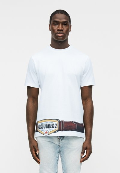 Άνδρας που φοράει λευκό T-shirt με εκτυπωμένη καφέ ζώνη και το λογότυπο "DSQUARED2" κοντά στο τελείωμα, συνδυασμένο με ανοιχτόχρωμα μπλε τζιν.
