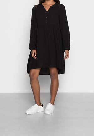 Frau trägt ein lockeres schwarzes, langärmliges, geknöpftes Kleid, kombiniert mit weißen flachen Sneakern, steht auf einem grauen Boden vor einer schlichten Wand.