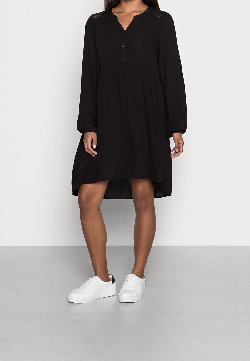 Femme portant une robe noire ample à manches longues boutonnée, associée à des baskets blanches basses, debout sur un sol gris contre un mur uni.