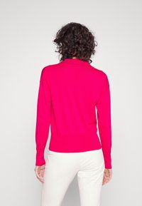 Pull en maille rose ajusté avec un col à revers, des manches longues et des poignets côtelés, présenté sur un fond neutre.