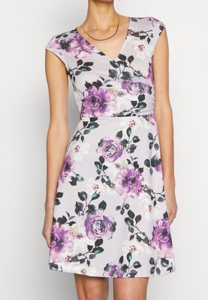 Kobieta w sukience na ramiączkach, z V-kształtnym dekoltem, sięgającej kolan, w fioletowo-biały wzór kwiatowy, z złotym łańcuszkiem na szyi.