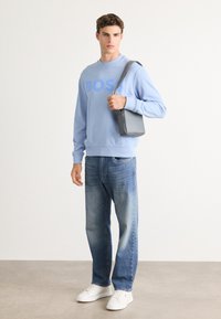 Joven wearing a light blue BOSS sweatshirt, blue jeans, white sneakers, holding a gray shoulder bag, standing against a white wall.