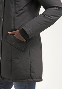 Osoba nosící černou prošívanou bundu s klopovým kapsou a patentním knoflíkem, ruka částečně v kapse, viditelná nad modrými džínami.