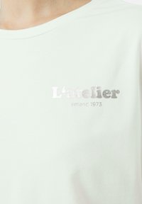 Hellgrünes T-Shirt mit weicher Textur und silberner gestickter Aufschrift "L'atelier alemand 1973" auf der Brust. Rundhalsausschnitt.