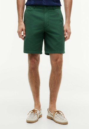 Uomo che indossa pantaloncini verde scuro fino al ginocchio, maglietta blu navy e scarpe casual bianche con lacci marroni, in piedi su sfondo bianco.