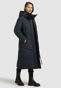 Lang, mørkeblå jakke med dunfyld og hætte, store kviltede sektioner, sidelommer med lynlås og trykknaplukning i kanten. Båret med sorte støvler.