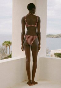Bikini a due pezzi di color bordeaux con strisce bianche, con top bandeau e parte inferiore abbinata, visto da dietro in un contesto costiero.