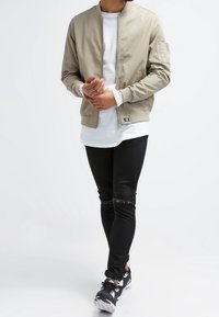 Man draagt een beige bomberjack over een wit shirt met lange mouwen, zwarte gescheurde skinny jeans en zwarte sneakers, loopt naar voren.