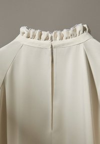 Vêtement couleur crème avec un décolleté volanté et une ouverture centrale dans le dos. Le tissu est lisse et léger, mettant en valeur un design minimaliste.