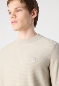 Beige neulotussa neulepaidassa on ribbitekstuuri ja olkasauman yksityiskohta, ja edessä on pieni neliönmuotoinen logo, jossa lukee "BOSS".
