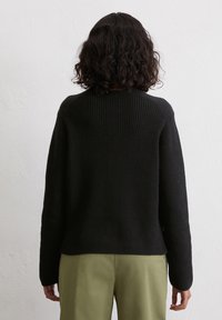 Schwarzer gestrickter Pullover mit geripptem Muster, tiefen Schultern und gerader Silhouette. Kombiniert mit olivgrünen Hosen.