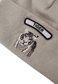 Berretto in maglia grigia con un ricamo di una tigre e un'etichetta bianca con la scritta "TIGER" in nero. Il tessuto ha un aspetto strutturato.