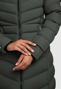 Dunkelgrüne Steppjacke mit vertikalem Reißverschluss, gesteppter Textur und Manschetten-Details. Die Hände sind vorne an der Jacke positioniert.