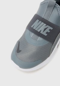 Pilkas sportinis batelis su tinkliniu viršumi, elastine juosta su "NIKE" prekių ženklu ir tekstūruotu padu. Papuoštas juodu šypsena logotipu.