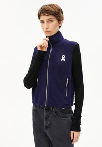 Marineblaues Fleece-Weste mit hohem Kragen, zwei vorderen Reißverschlusstaschen und einem weißen Bandakzent. Trägt man über einem schwarzen Langarmoberteil.
