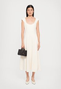 Ein enganliegendes, cremefarbenes Kleid mit kurzen Ärmeln und einem runden Ausschnitt, kombiniert mit einer kleinen schwarzen Handtasche und spitzen weißen Ballerinas.