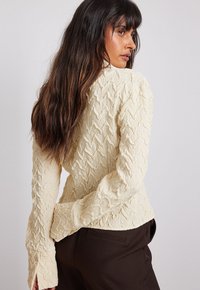 Giacca a maniche lunghe in crema texturizzata con un motivo ondulato, design aderente e polsini aperti, abbinata a pantaloni scuri.
