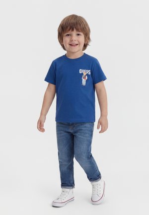 Uśmiechnięty młody chłopiec stoi, ubrany w niebieski T-shirt z rysunkiem i napisem „GUESS”, niebieskie dżinsy oraz białe trampki na jednolitym tle.