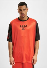 Rød og sort kortærmet skjorte med et logo, der står "ECKŌ unlimited." Skjorten har en glat tekstur og en løs pasform. Den har kontrastfarvede ærmer.