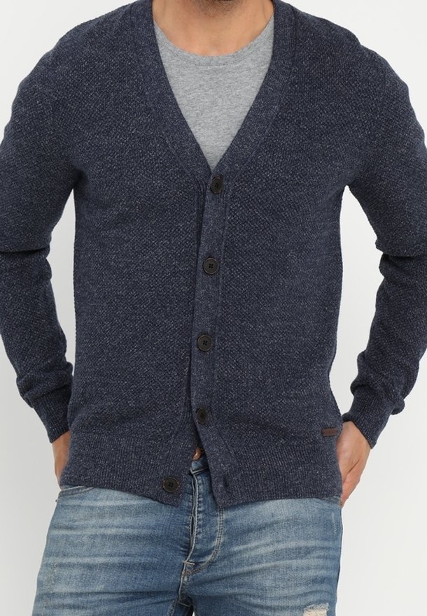 Dunkelblaue Strickjacke mit V-Ausschnitt, sechs Knöpfen und gerippten Bündchen. Getragen über einem grauen Shirt, kombiniert mit hellblauen, verwaschenen Jeans.