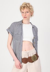 Striped short-sleeve shirt with front pocket, paired with white cropped top and beige textured pants, accented by a wide brown belt with circular details.