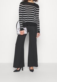 Sweter w paski czarno-biały połączony z czarnymi spodniami o szerokich nogawkach i czarnymi szpilkami, trzymając małą srebrną torebkę metaliczną.