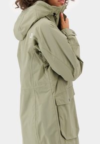 Veste imperméable vert olive clair avec un col montant, une capuche ajustable et des poches avant. Texture lisse et coupe décontractée.