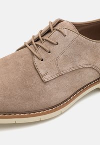 Zapato de vestir de ante beige con punta redonda, que presenta un diseño de cordones, suela crema y costuras contrastantes a lo largo del borde.