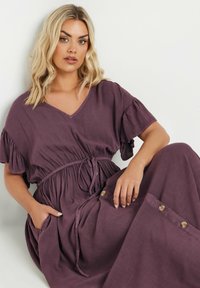 Lila, flödande maxikjol gjord av mjukt tyg, med V-hals, volangärmar, dragsko i midjan, sidofickor och dekorativa knappar.