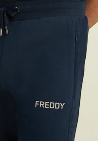 Joggers azul marino de tela suave, con cintura ajustable con cordón, bolsillos laterales y texto plateado "FREDDY" en el muslo izquierdo.