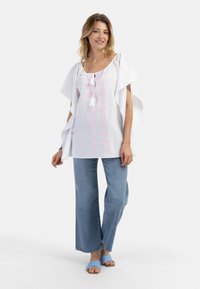 Top in cotone bianco con motivi floreali ricamati in rosa, caratterizzato da un colletto con cordino e maniche ampie, abbinato a jeans a gamba larga blu e sandali blu.