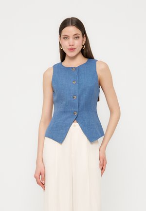 Frau trägt ärmelloses, blaues, geknöpftes Oberteil mit spitz zulaufendem Saum, kombiniert mit weißen, hoch geschnittenen, weit geschnittenen Hosen vor schlichtem Hintergrund.