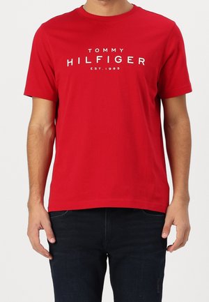 Hombre vistiendo una camiseta roja de manga corta con el texto blanco "Tommy Hilfiger EST. 1985", combinado con jeans oscuros.