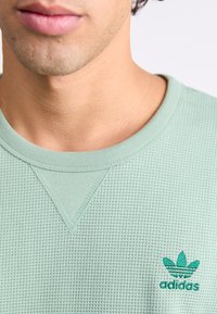 Šviesiai žalias tekstūruotas nadažas su apykakle. Kairėje krūtinės pusėje yra išsiuvinėtas žalias Adidas logotipas.