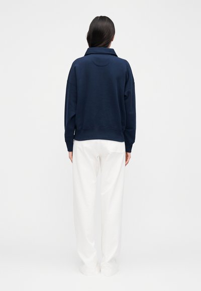 Γυναίκα με μακριά σκούρα μαλλιά, που φοράει μπλε σκούρο μπλουζάκι με μακριά μανίκια και άνετο λευκό παντελόνι, στέκεται γυρισμένη με την πλάτη προς ένα απλό φόντο.