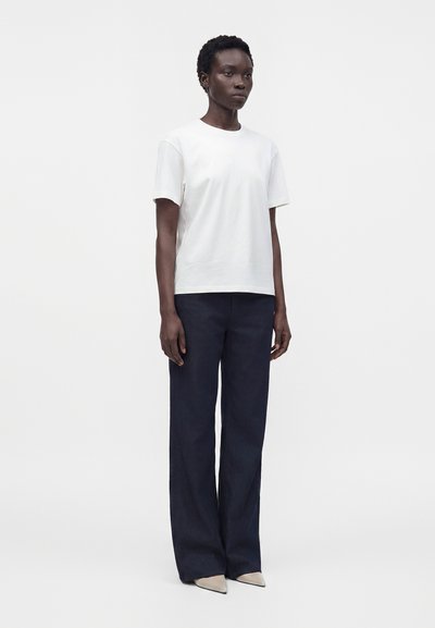 Standing person wearing a plain white short-sleeve shirt, loose dark pants, and light-colored pointed shoes against a white background.