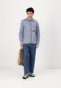 Lys blå jakke med glidelås, stor lomme på brystet og sidelommer med glidelås. Kombinert med vide blå jeans og grå joggesko.