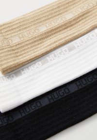 Tri páry rebrovaných ponožiek v béžovej, bielej a čiernej farbe; každé má na sebe logo stuhu s nápisom "HUGO" v jemnom vzore.