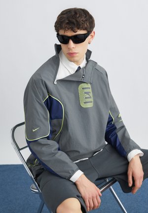 Ung mand iført mørke solbriller, grå og marineblå vindjakke med limefarvede detaljer, hvid skjorte med krave og grå shorts, der sidder på en metalstol.