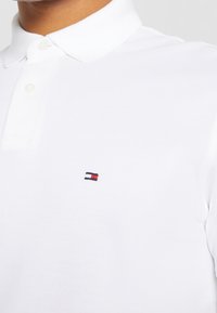 Polo blanc en tissu texturé, avec un col à trois boutons et un petit logo brodé en rouge, blanc et bleu.