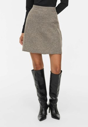 Jupe courte en maille beige portée avec des manches longues noires et des bottes en cuir noires montantes jusqu'aux genoux avec des bouts pointus et des talons.