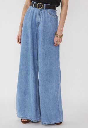 Jean denim bleu clair à jambes larges avec des plis, porté avec une ceinture noire et des bijoux dorés à la main droite.